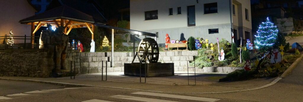Night scene of a small town Christmas display with a wooden gazebo, statues, a water wheel centerpiece, and a lit Christmas tree by a stone wall garden orna ments outside a house.