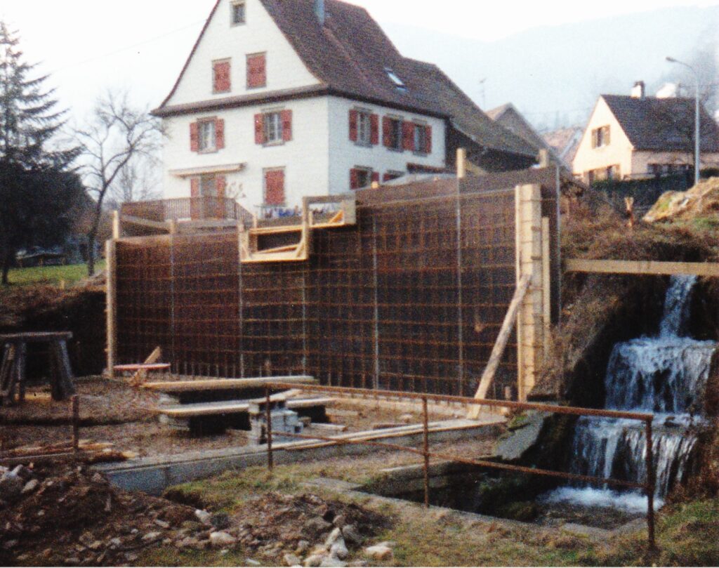 Construction site with a tall wooden formwork wall next to a small waterfall, houses visible in the background.