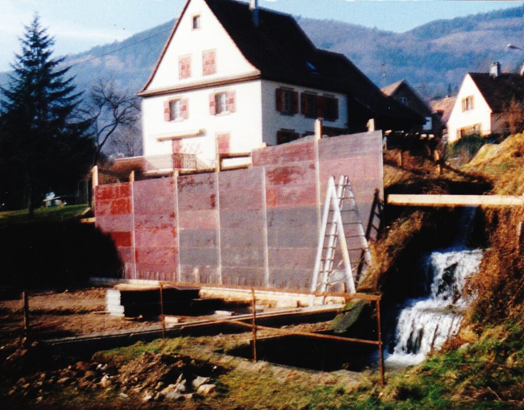 Construction site with pink-gray barrier panels, a ladder, and a small waterfall in the foreground against a hillside neighborhood building behind it