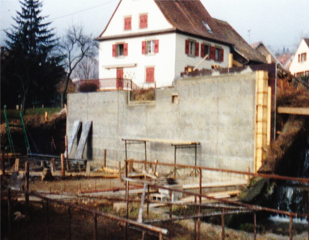 Construction site with a tall concrete wall, scaffolding, and building materials; a white house with red shutters behind it.