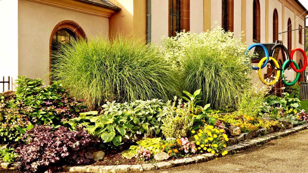 Garden with tall grasses and colorful flowers along a stone border beside a beige building with arched windows; Olympic rings sculpture on the right.