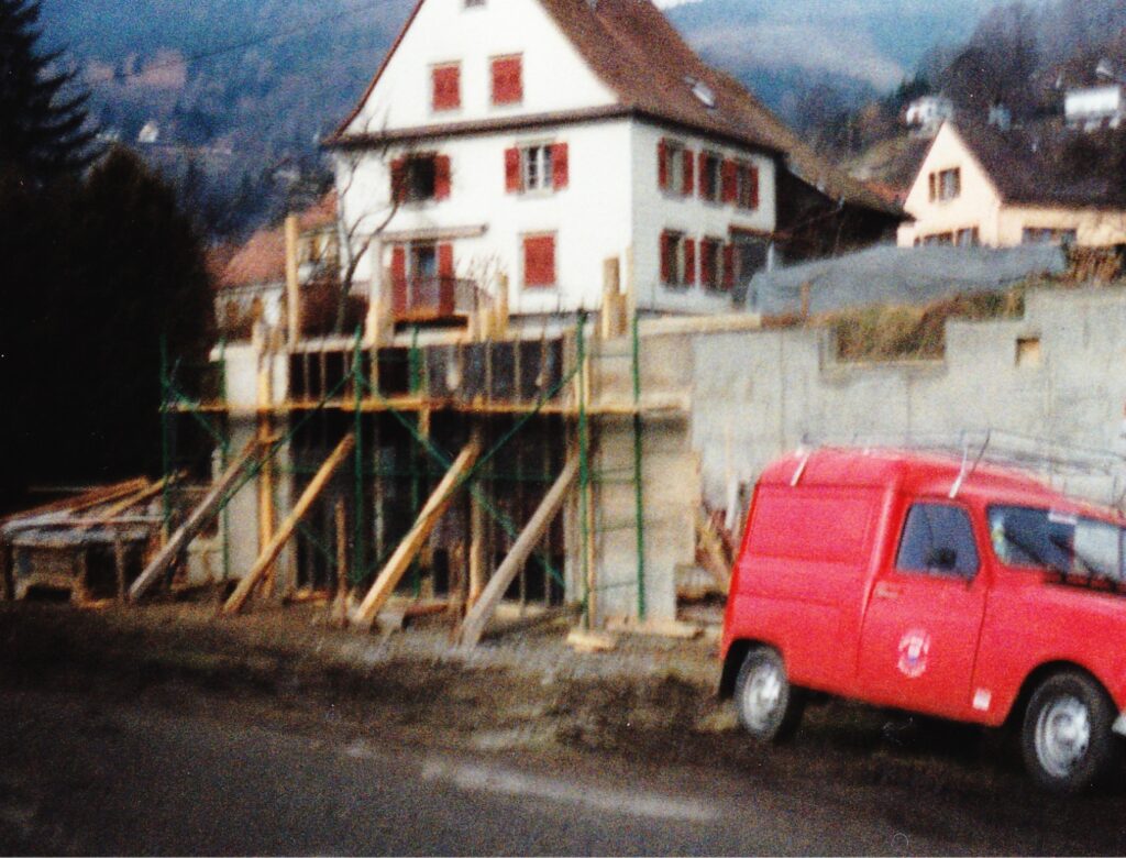 Construction site at a hill with wooden scaffolding and concrete forms, white house in background, red van parked nearby.