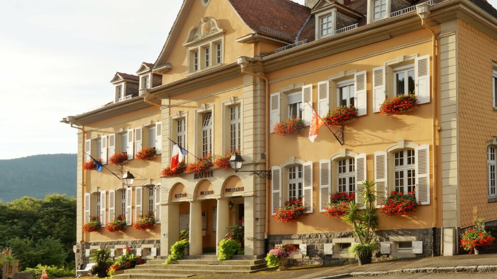 Sunlit yellow town hall with white shutters and red flower boxes along the façade