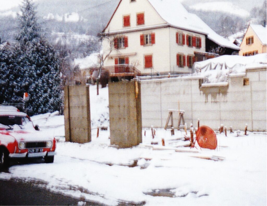 Snowy construction site with two tall concrete columns, a red car parked at left, and a large house with red shutters in the background.