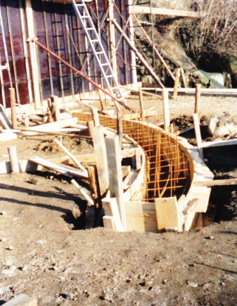 Construction site with curved wooden formwork and orange metal rebar for a circular concrete wall, ladder leaning against a framed building in the background.