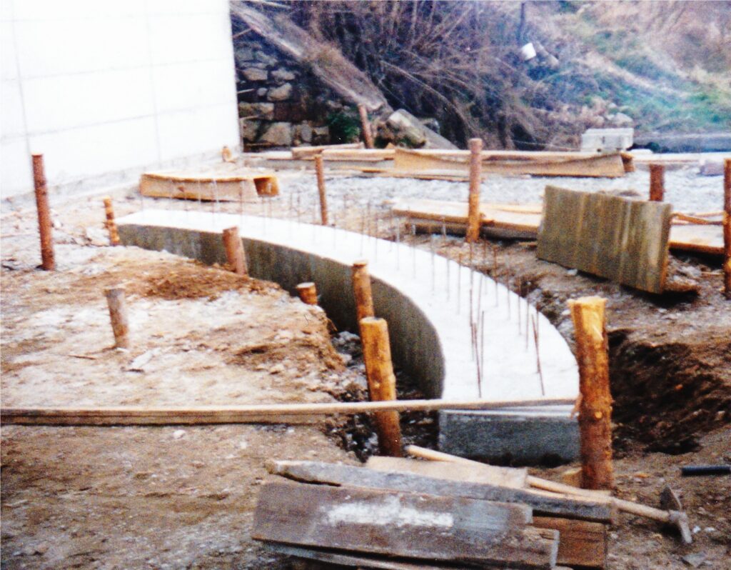 Curved concrete footing under construction with exposed rebar and wooden stakes at a muddy site, hillside in background