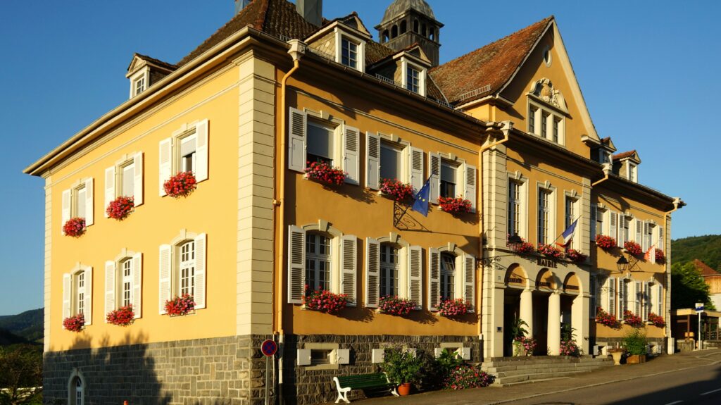 Yellow European-style building with white shutters, flower boxes along windows, and flags under a clear blue sky.
