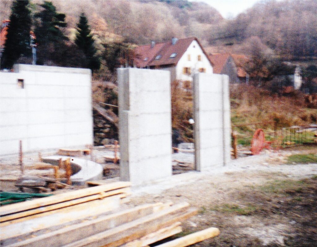 Construction site with tall concrete block walls, lumber, and a curved concrete form in the foreground; a white house with a red roof sits in the background.