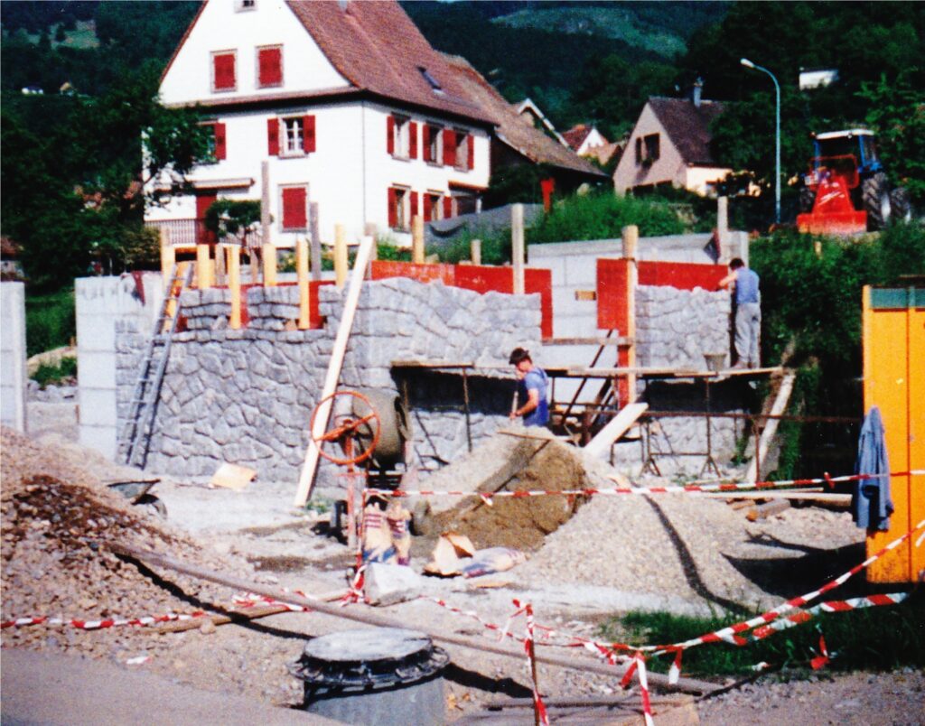 Construction site with stone foundation walls, scaffolding, and workers pouring gravel in a suburban area with houses in the background