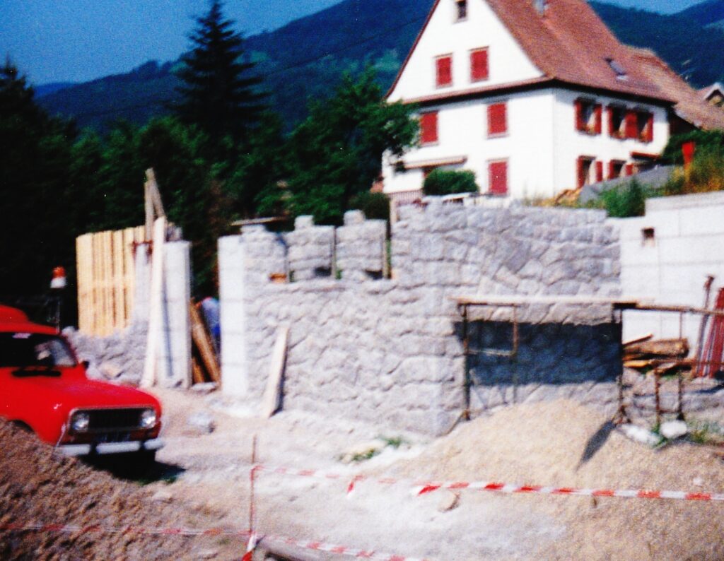 Construction site with a partially built stone wall, gravel ground, and a red car in the foreground; a white house with red shutters behind.