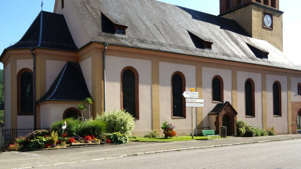 Facade of a beige church with arched stained-glass windows, slate roof, and a clock tower in the background, with a flower garden in front.