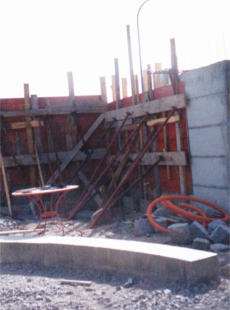 Construction site with wooden formwork, vertical stakes, and scattered rubble near a curved concrete wall.