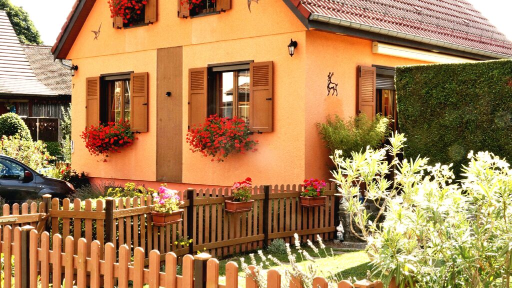 Orange stucco house with brown shutters and window boxes filled with red flowers, a wooden picket fence, and a lush garden in front.
