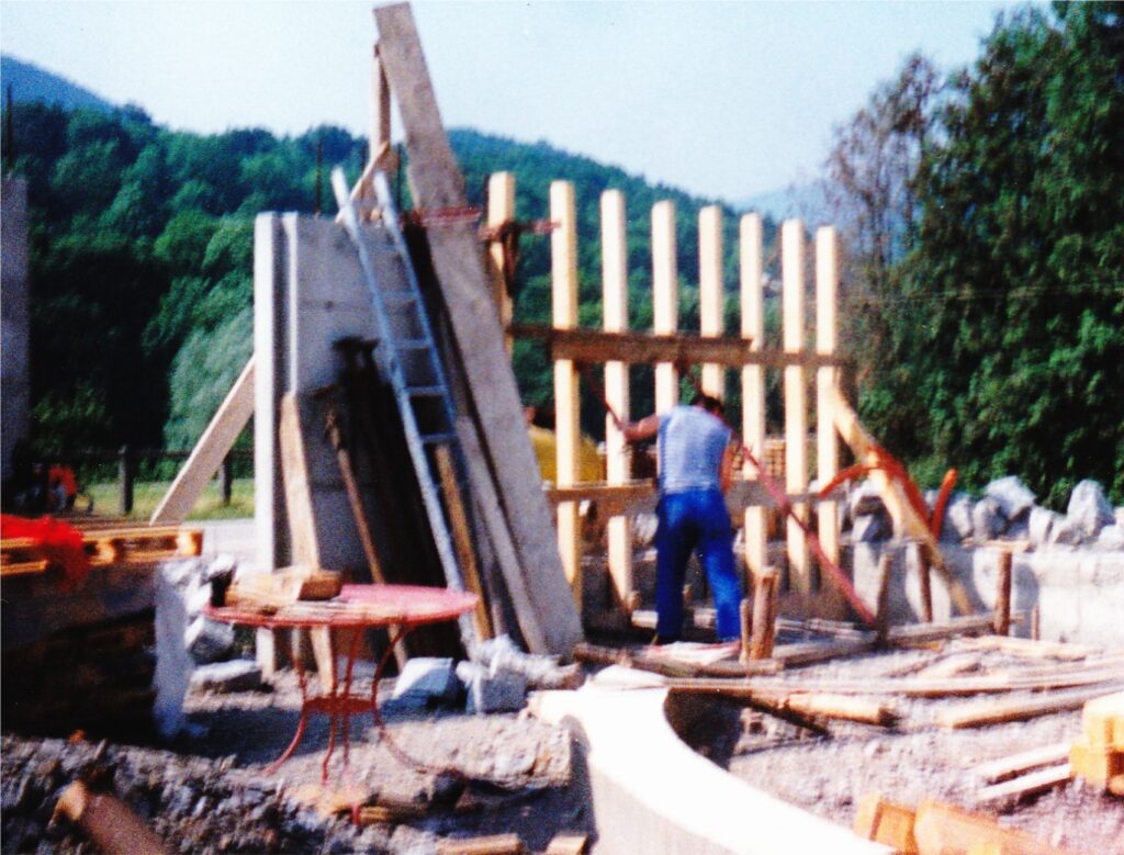 Construction site with a worker building a wooden frame for a wall; lumber, concrete blocks, and tools scattered around outdoors.
