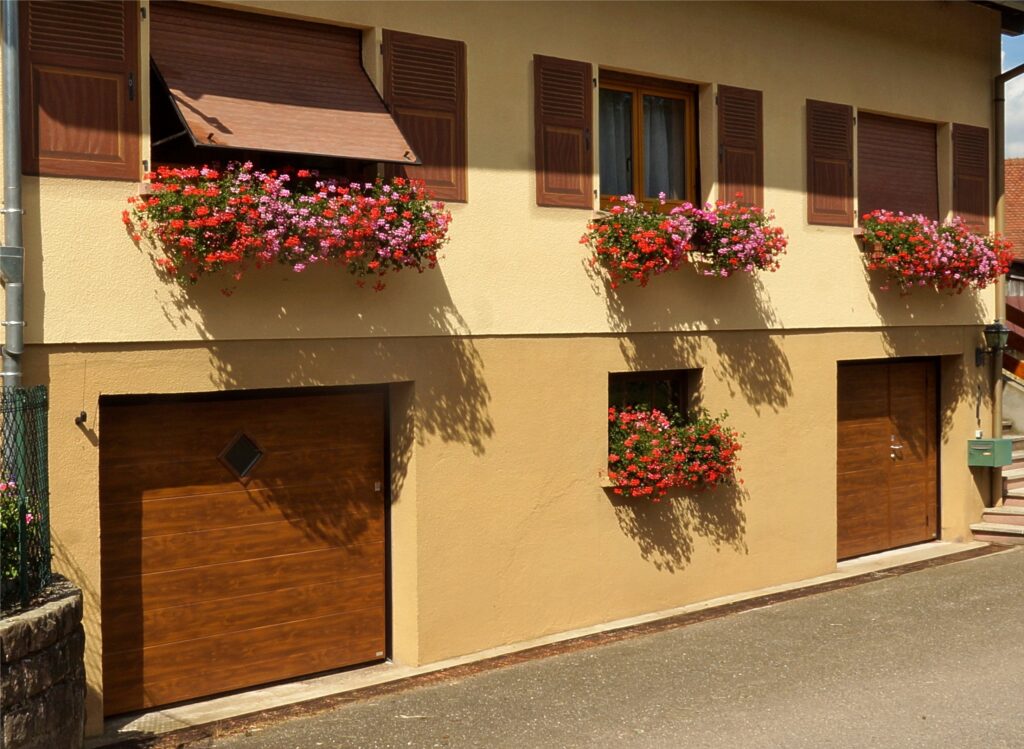 Beige building facade with brown wooden shutters, window boxes filled with red and pink flowers, and closed garage doors.