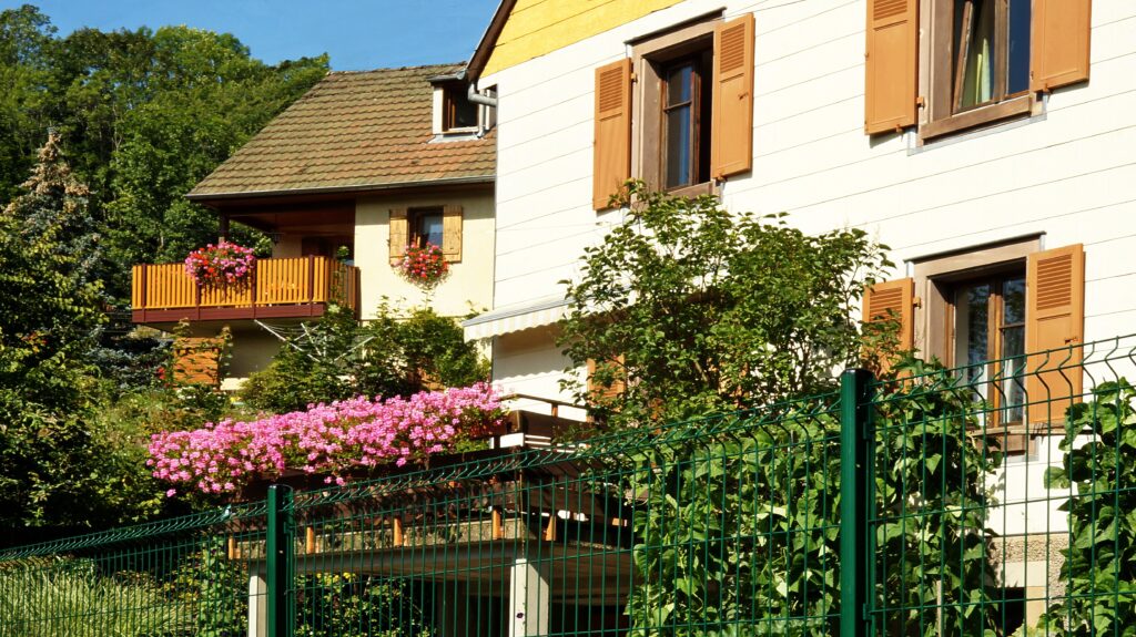 Two-story house with beige walls, brown shutters, and a wooden balcony filled with pink flowers, surrounded by green garden and a green fence.