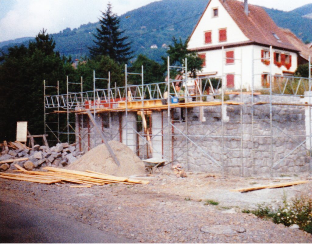 Scaffolding surrounds a partially built stone wall at a construction site in front of a white house with red shutters against a hillside backdrop.