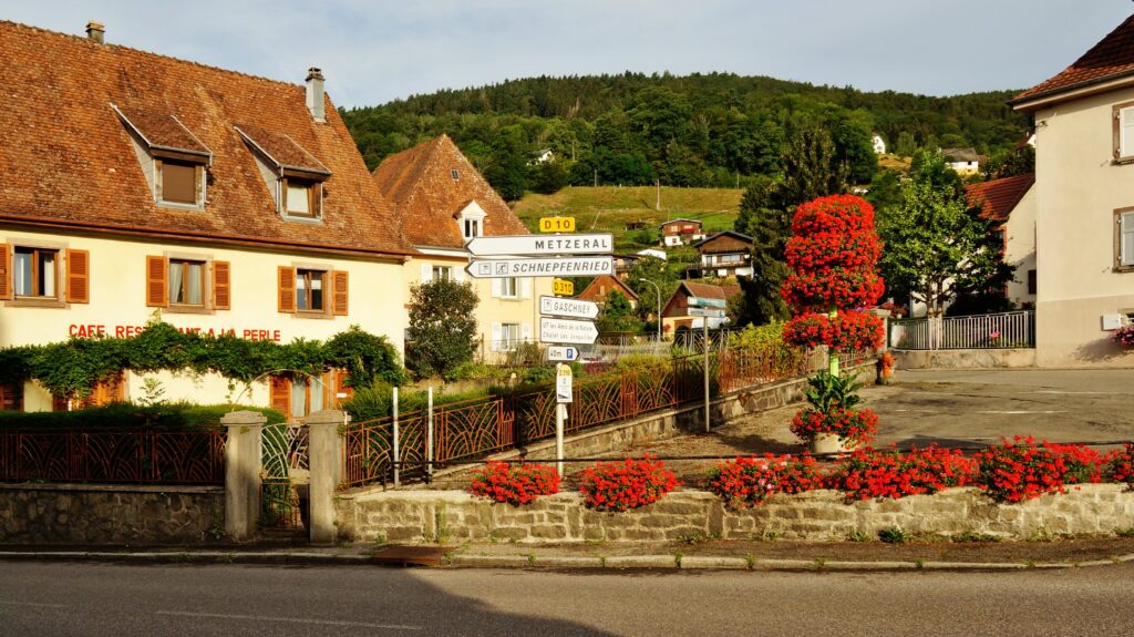 Sunny village street with red flowering planters along a stone wall and pastel houses in the background.