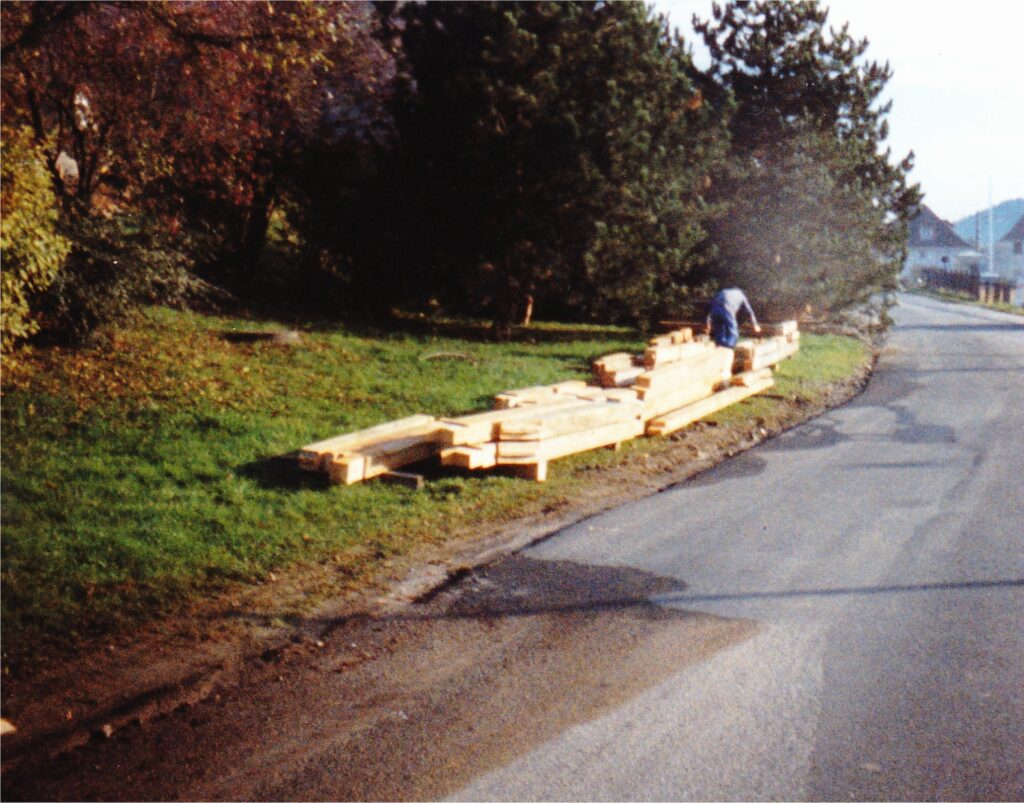 Person stacking long wooden planks along the roadside with trees in the background, in a residential area.
