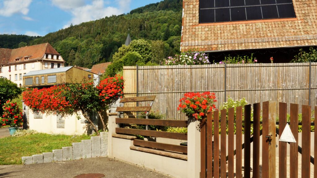 Residential backyard with a wooden slat fence, red flowering bushes, and houses on a hillside under a blue sky, sunny day.