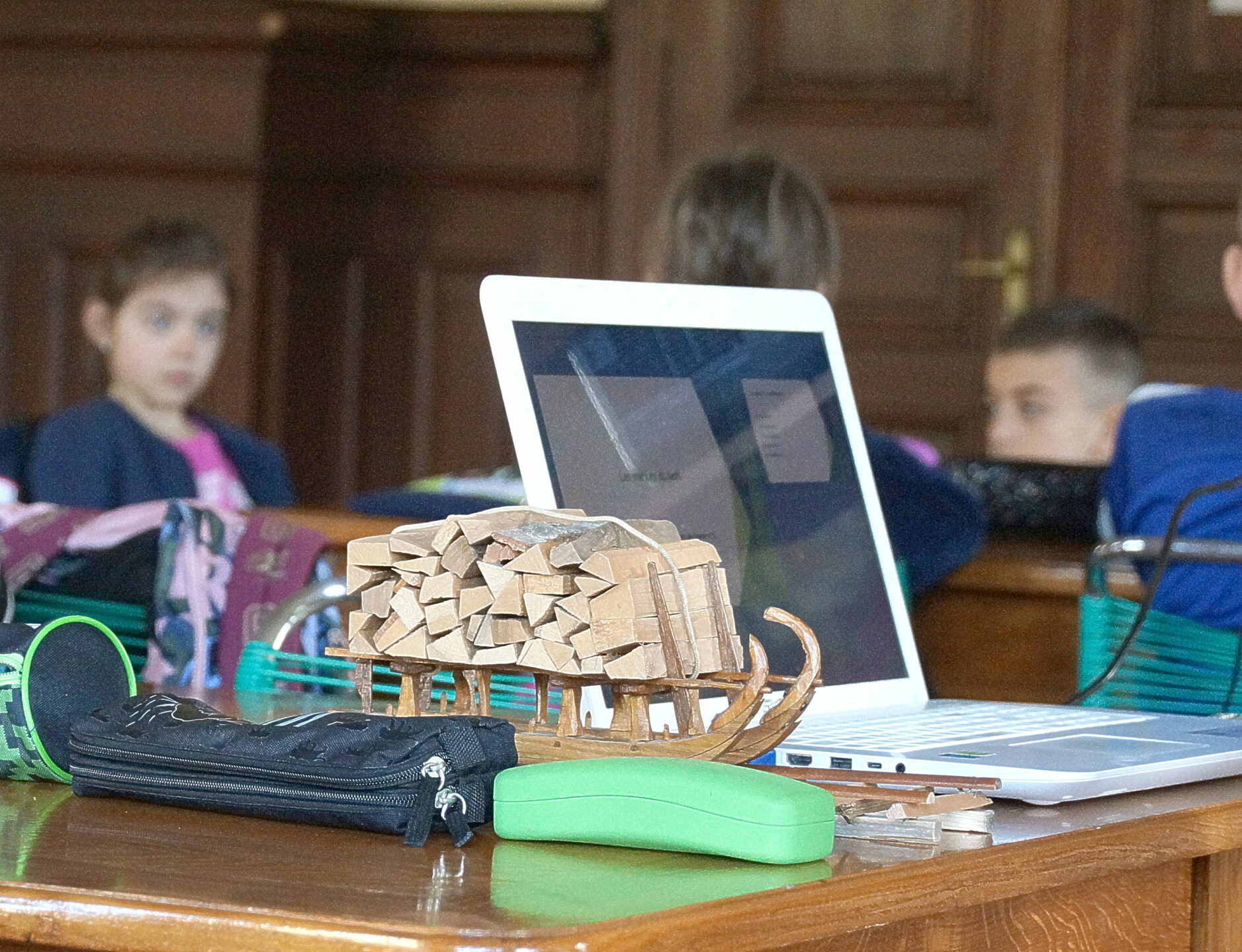 Children sit at a wooden table with backpacks and a white laptop; a cardboard sculpture sits in the foreground, suggesting a classroom or workshop setting.