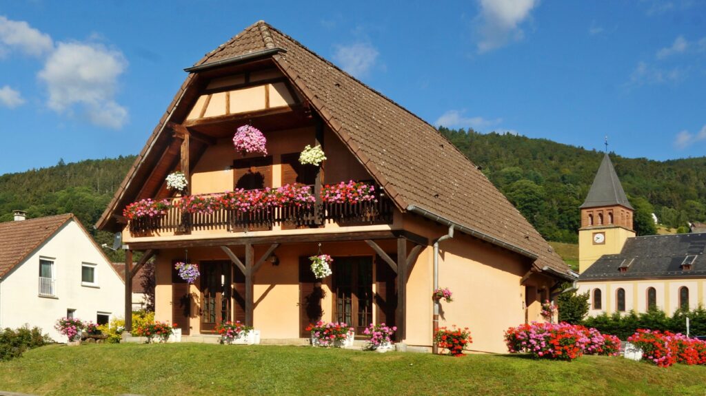 Peach-colored chalet with wooden balcony overflowing with pink, red, and white flowers under a blue sky.