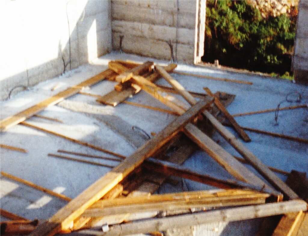 Wooden planks and scraps scattered on a concrete floor inside a partially built structure with cinder block walls and an open doorway to trees outside