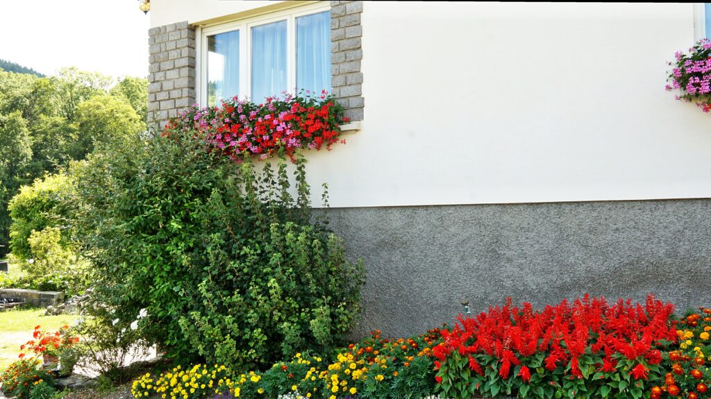 Exterior of a house with a flower-filled window box, stone posts, and a lush garden bed of yellow and red flowers along a white textured wall.