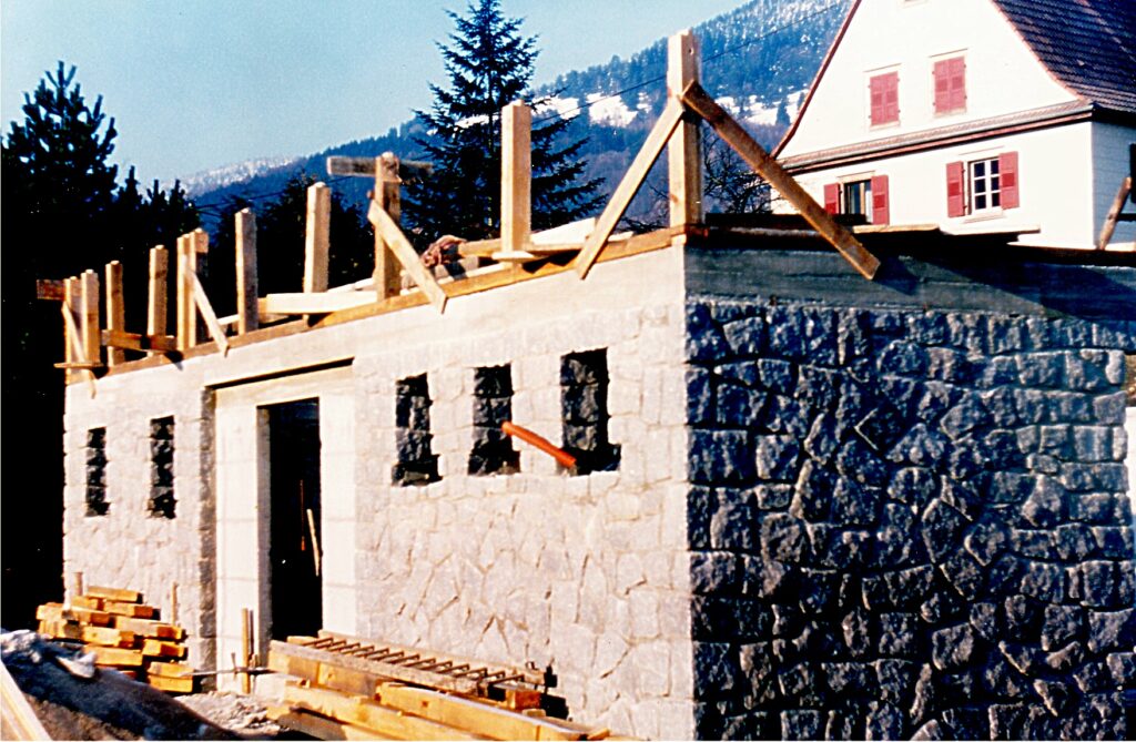Stone-and-block building under construction with wooden scaffolding on top of a stone wall, mountain backdrop in a rural setting