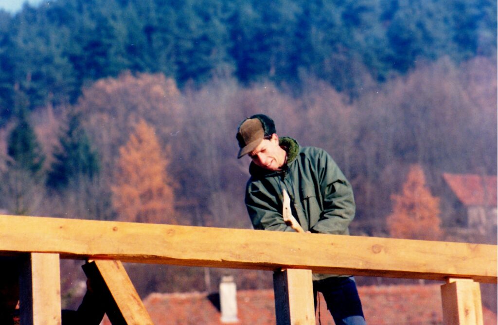 Man in a green jacket and brown cap leaning on a wooden railing outdoors, with a forested hillside in the background.