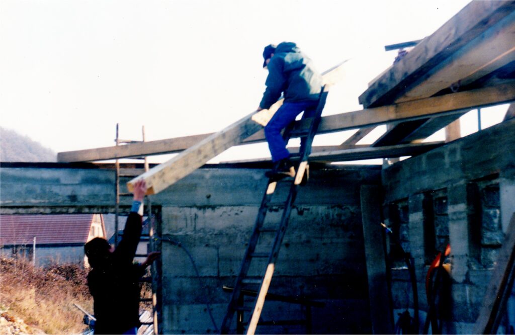 Two construction workers coordinate on a wooden scaffold, passing a beam on an unfinished building exterior, with one on a ladder.