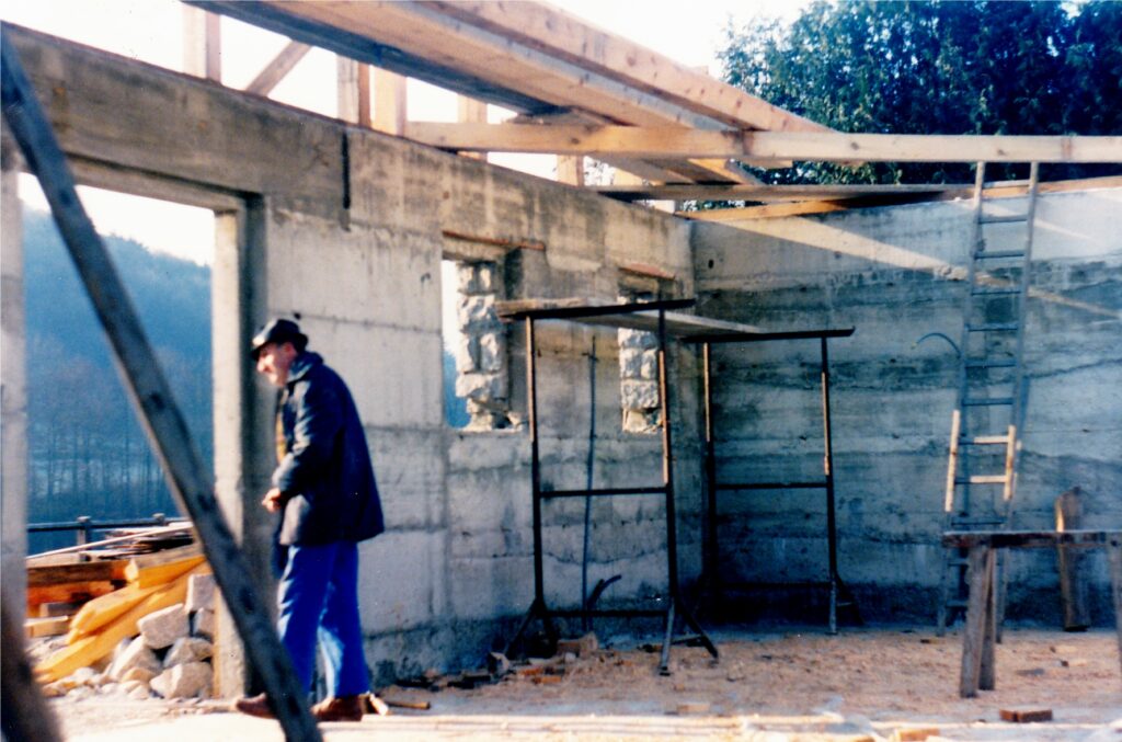 Construction worker in a blue jacket and hat stands beside an unfinished concrete wall with wooden beams overhead.