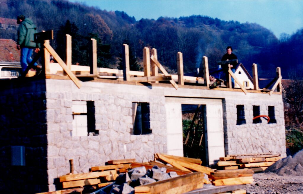 Stone building under construction with wooden framing on the roof, two workers standing on top, and piles of lumber in the foreground.