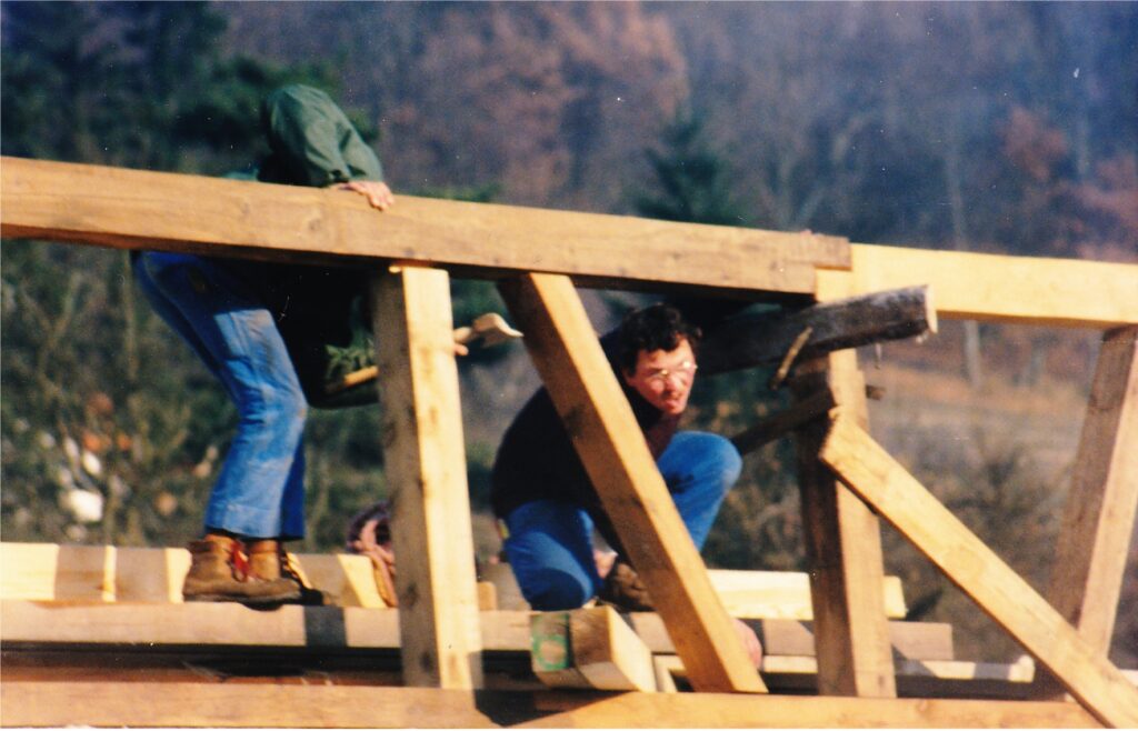 Two workers assemble a wooden frame outdoors, one crouched under a beam while another stands on the structure with boots visible.