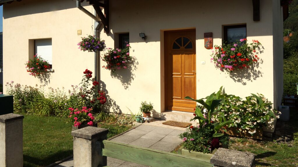 Cream-colored house with a wooden front door, two hanging flower baskets, and a flower-filled walkway garden.