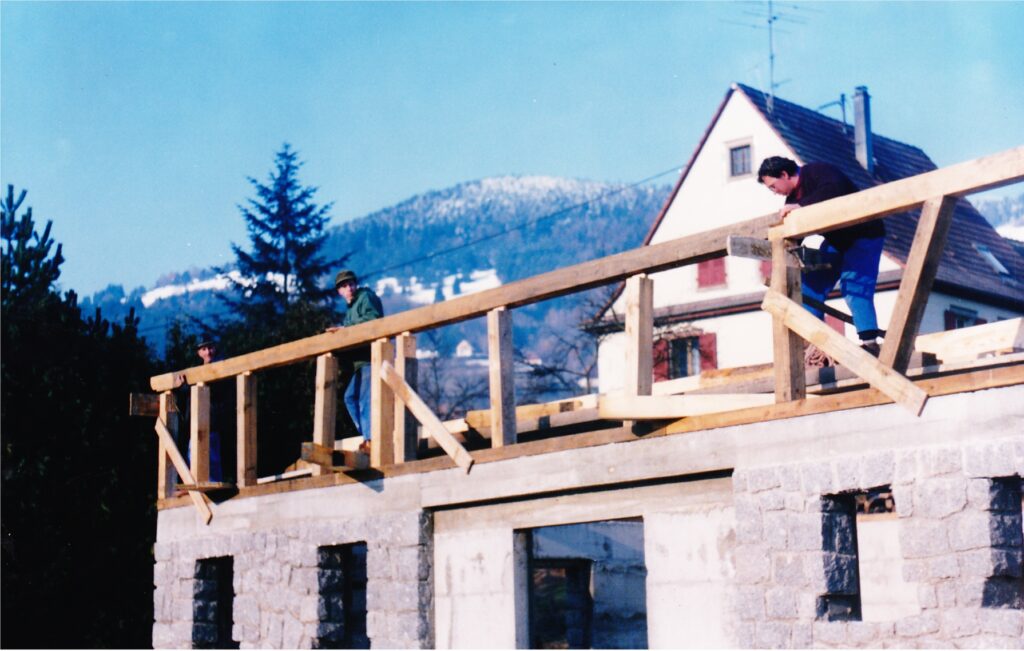 Three construction workers lean on a wooden railing atop a partially built stone building, with snow-capped hills in the blue sky background.