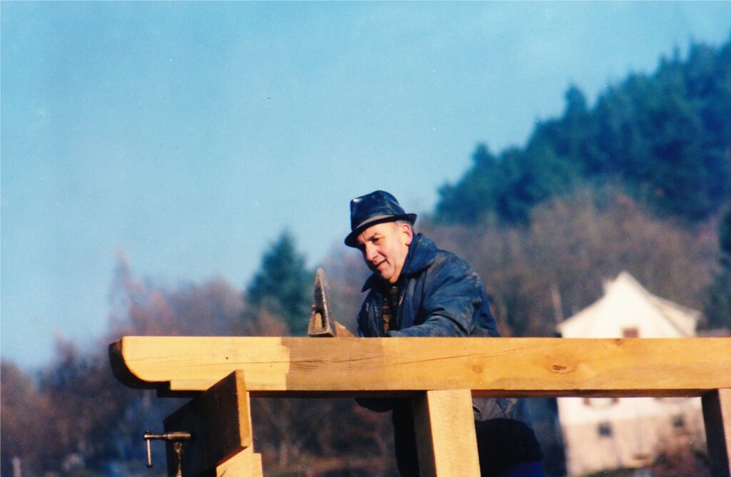 Man wearing a dark hat and blue jacket stands behind a wooden frame, working outdoors on a construction project.