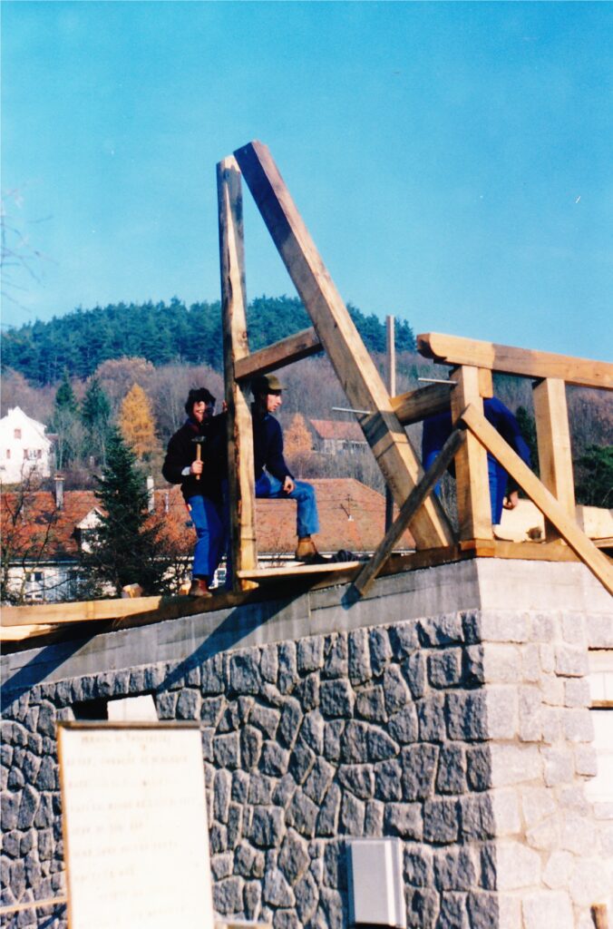Construction workers on a wooden frame atop a stone bridge against a clear blue sky, mid-construction.