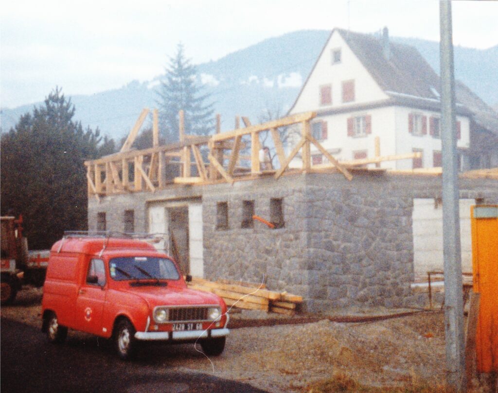 Red vintage van parked in front of a stone building under construction with wooden roof framing, hills and a house in the background