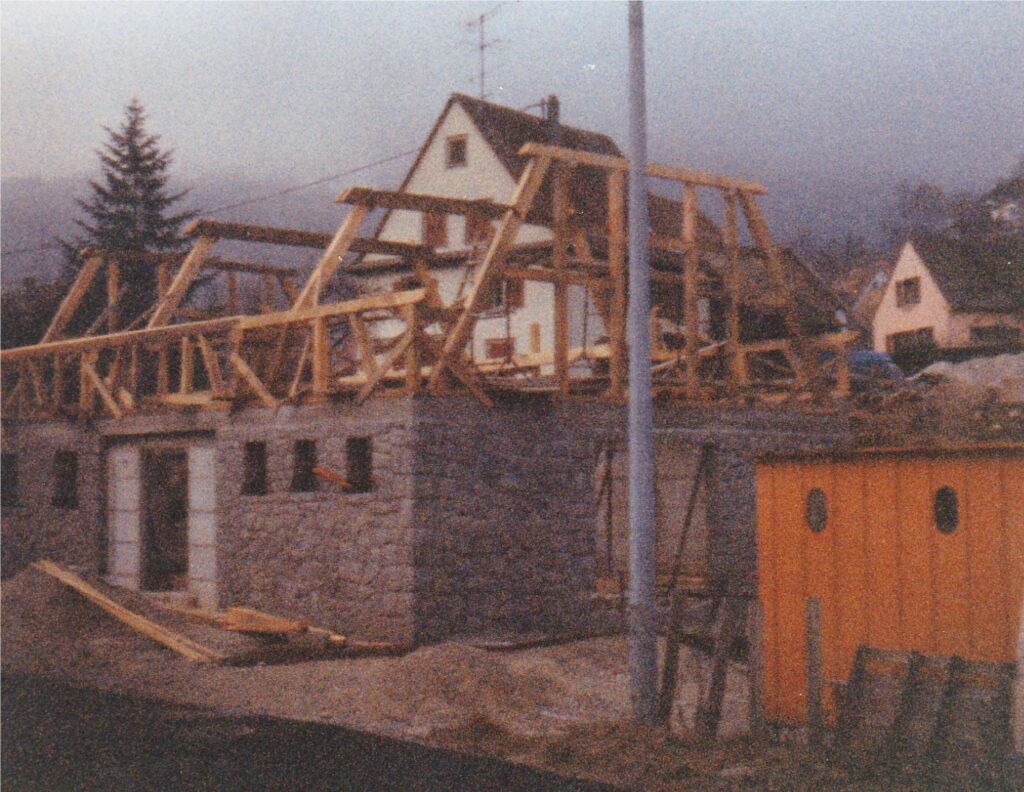 Wooden roof framing being added to a stone building, with an orange shed nearby and houses in the background at dusk