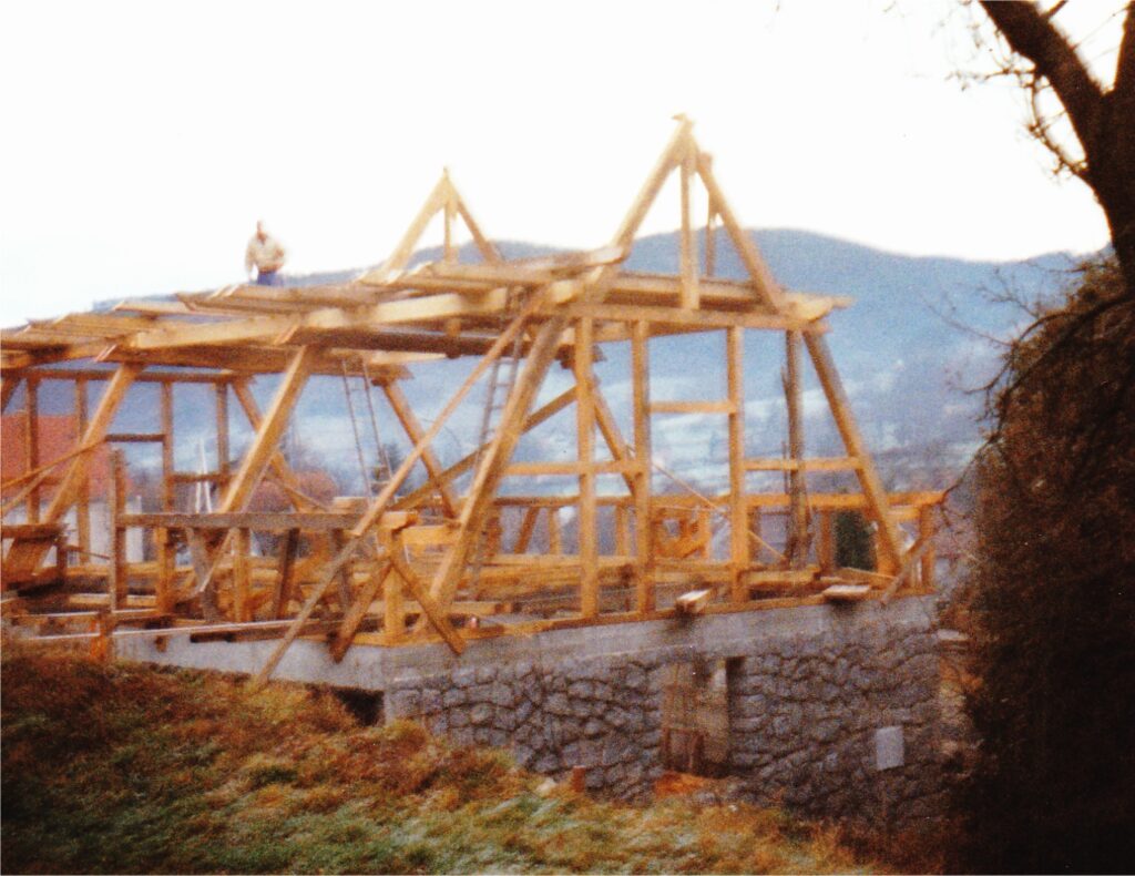 Wood-framed house under construction on a stone foundation, with a person on the roof trusses and mountains behind.