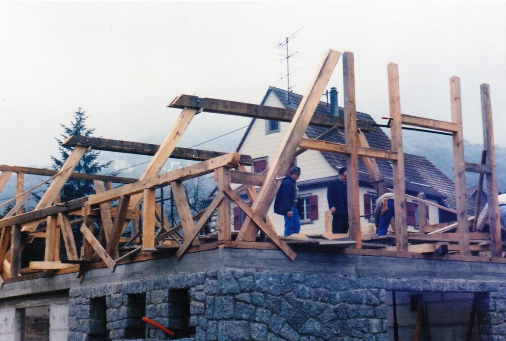 Wood framed building under construction with workers standing on a stone foundation, residential houses in the background.