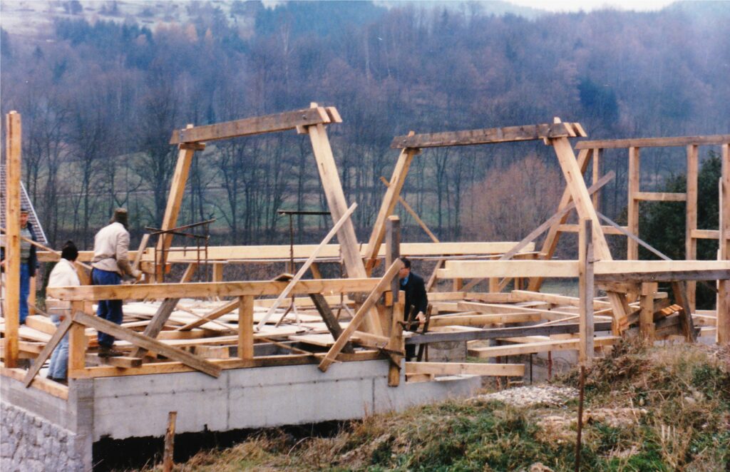 Wood-framed building under construction on a concrete foundation with workers handling lumber in a rural, hillside setting.