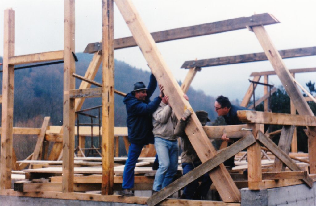 Men work together to lift a large wooden beam within a timber-framed building under construction outdoors, with hills in the background.