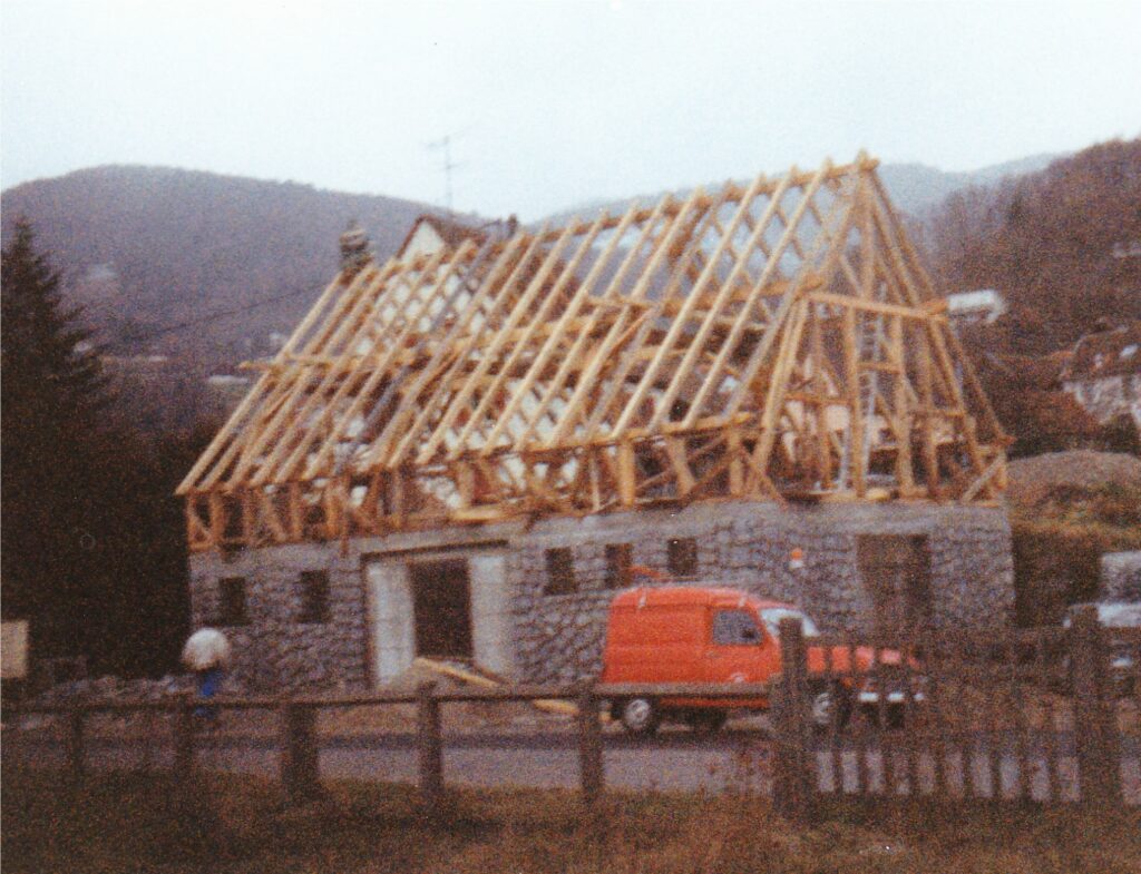 House under construction with wooden roof trusses over a stone masonry base; red van parked in front.
