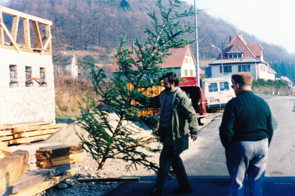 Two men walk along a street past a construction site with stacked lumber, a small evergreen tree, and houses in the background.