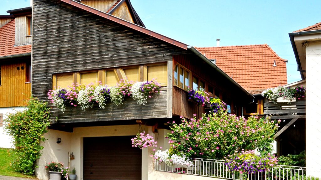 Two-story house with dark wooden siding, red-tiled roof, and window boxes of pink, white, and purple flowers under a blue sky.