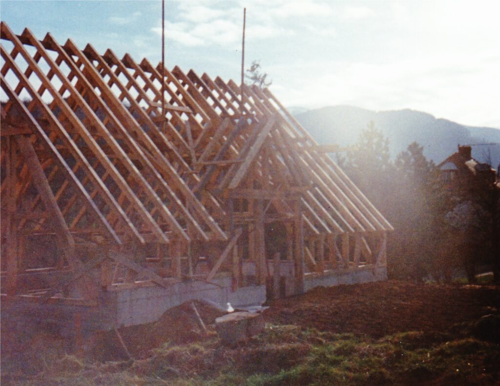 Wooden roof trusses forming a framed house under construction, set against hills and a partly cloudy sky.