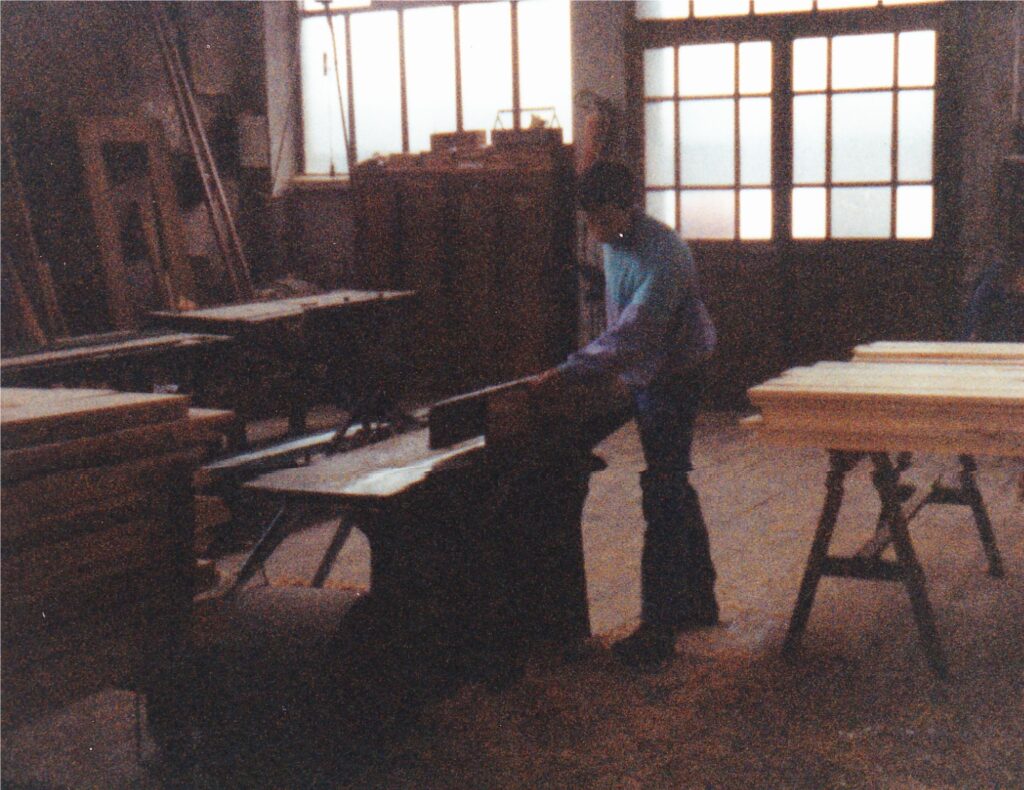 Man wearing a blue shirt works with a piece of wood on a table saw in a cluttered woodshop, with stacks of lumber in the background.