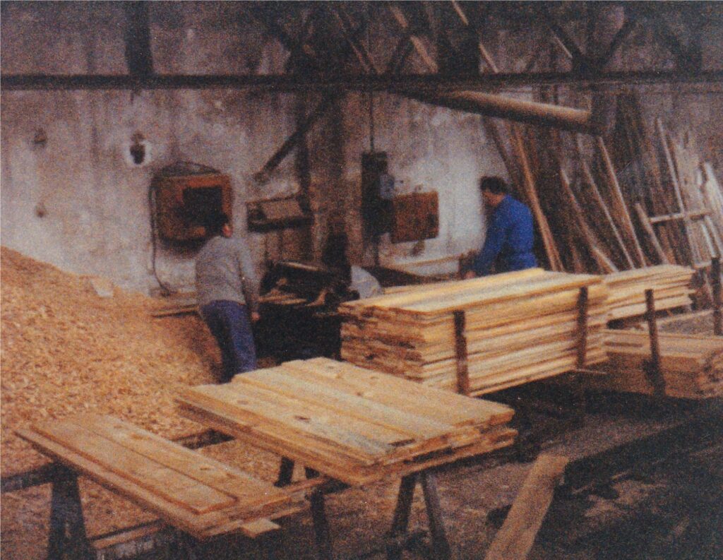 Two workers in a woodshop feed boards into a saw, surrounded by stacks of lumber and a dusty floor.